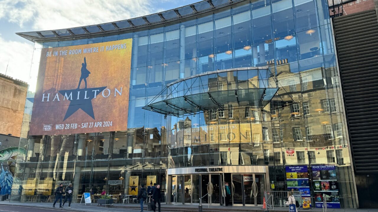Modern glass-fronted theater building with large "Hamilton" musical poster and reflection of surrounding cityscape, people walking in front.