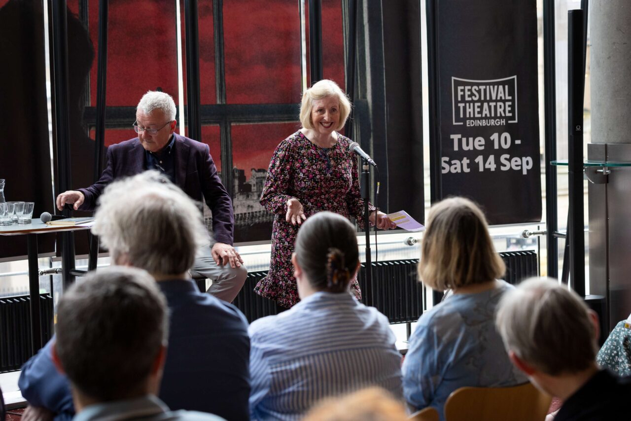 A woman speaks at a microphone to an audience during a daytime event, with a man seated beside her and a Festival Theatre Edinburgh banner in the background.