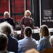 A woman speaks at a microphone to an audience during a daytime event, with a man seated beside her and a Festival Theatre Edinburgh banner in the background.