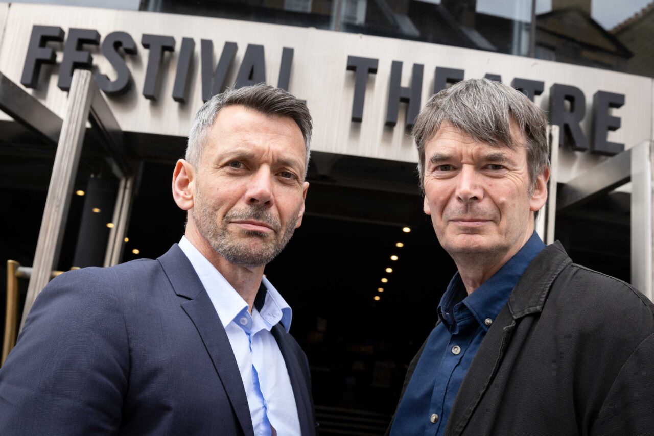 Two men stand in front of a building with a sign reading "Festival Theatre.