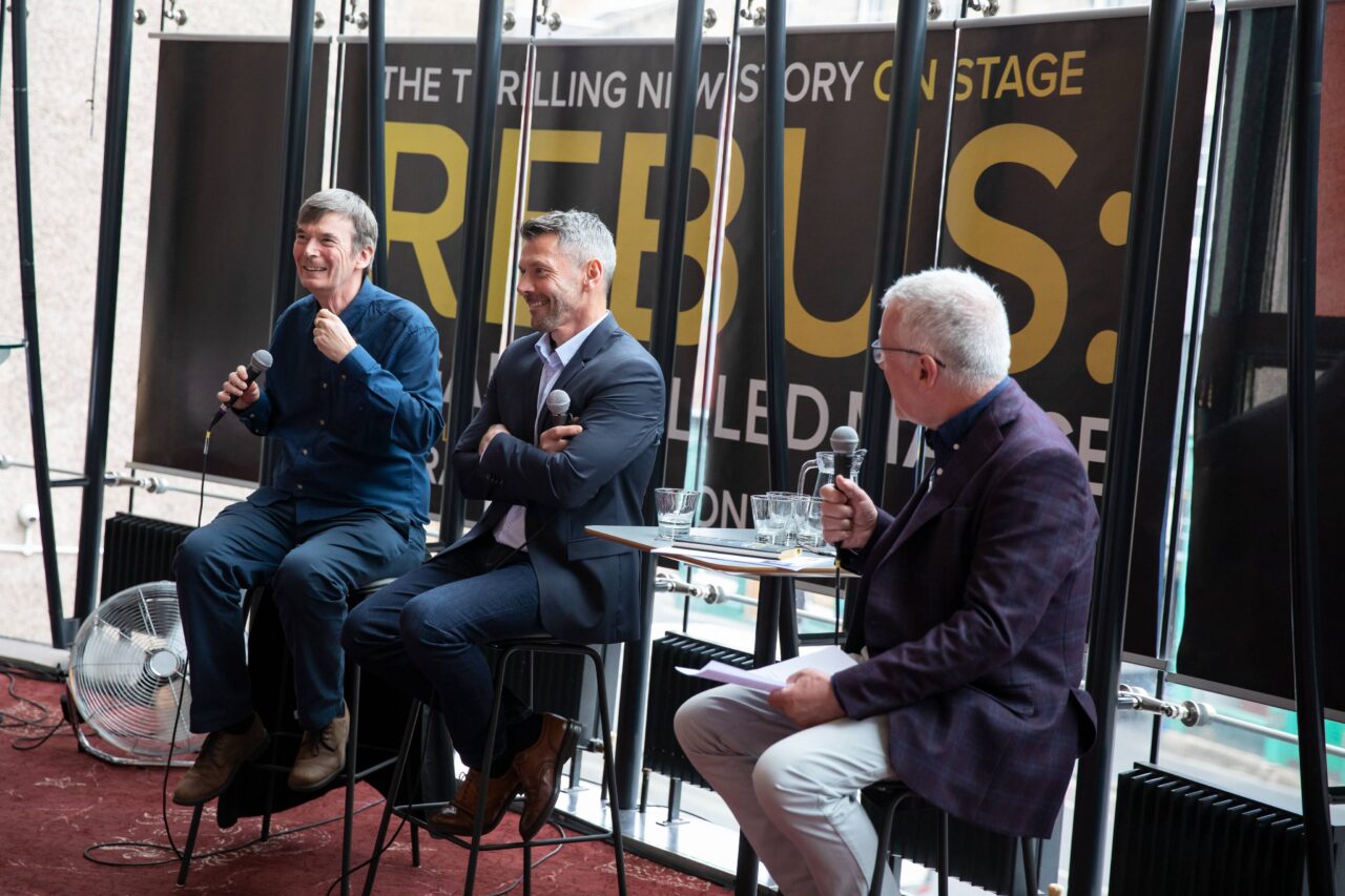 Three men sit on stools in front of a large REBUS stage sign, speaking into microphones during a panel discussion.