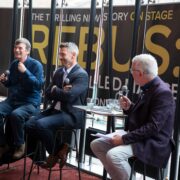 Three men sit on stools in front of a large REBUS stage sign, speaking into microphones during a panel discussion.