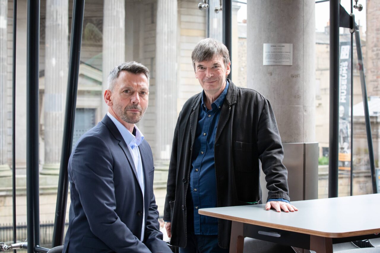 Two men are standing and sitting beside a table indoors, with stone columns and large windows visible in the background.