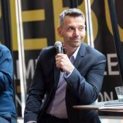 A man in a suit sits at a table holding a microphone, with another man seated beside him; glasses and papers are on the table.