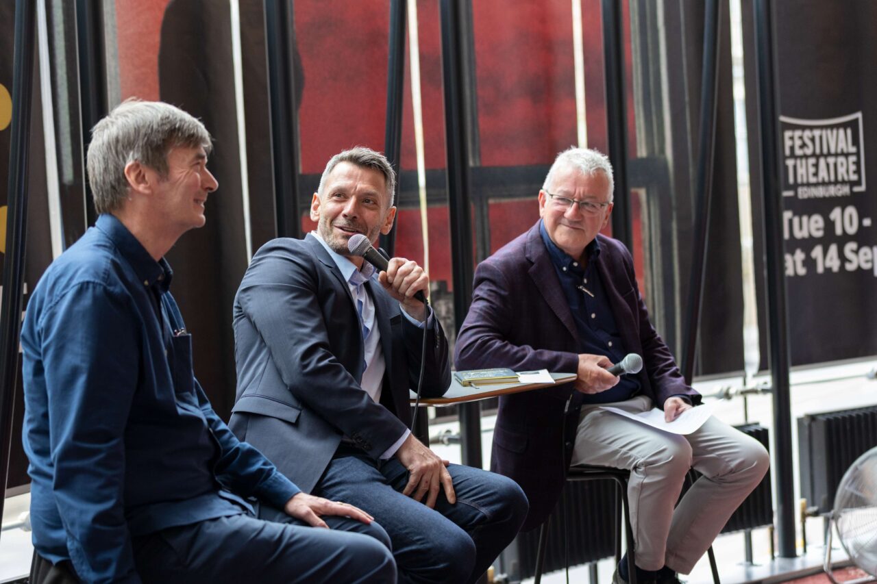 Three men sit on stools holding microphones, engaged in conversation at an indoor event, with Festival Theatre Edinburgh banners visible in the background.