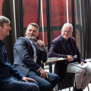 Three men sit on stools holding microphones, engaged in conversation at an indoor event, with Festival Theatre Edinburgh banners visible in the background.