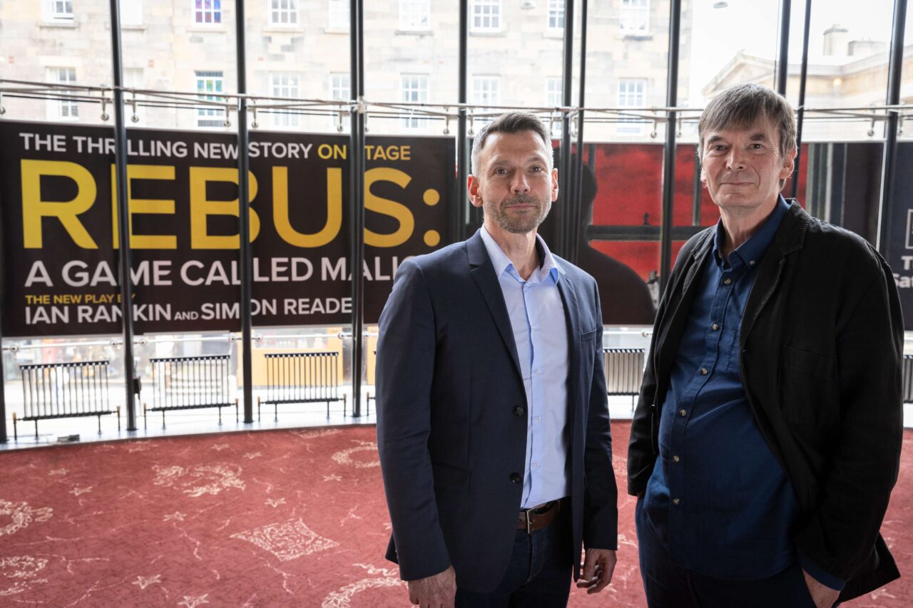 Two men in business-casual attire stand indoors in front of large promotional posters for "REBUS: A Game Called Malice.