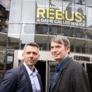 Two men standing outside the Festival Theatre, with a sign above them reading "Rebus: A Game Called Malice" by Ian Rankin.