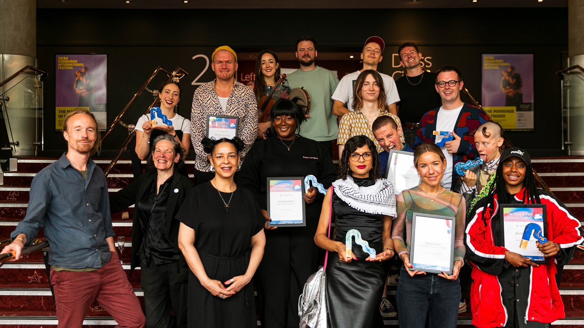 A group of people stand on stairs, holding certificates and blue dinosaur-shaped trophies, posing for a photo at an indoor event.