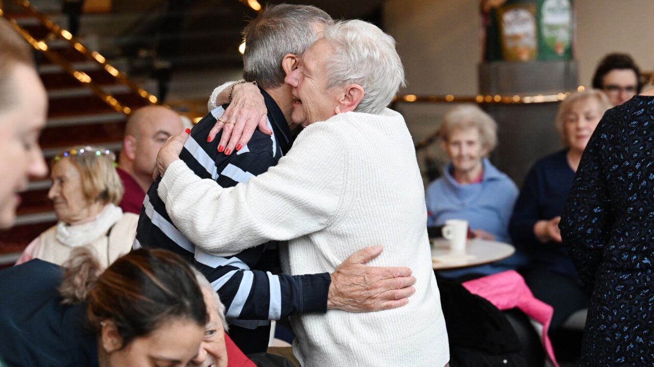 Two elderly people embrace in a hug at a social gathering, while others sit and watch in the background.