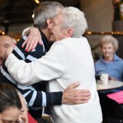 Two elderly people embrace in a hug at a social gathering, while others sit and watch in the background.
