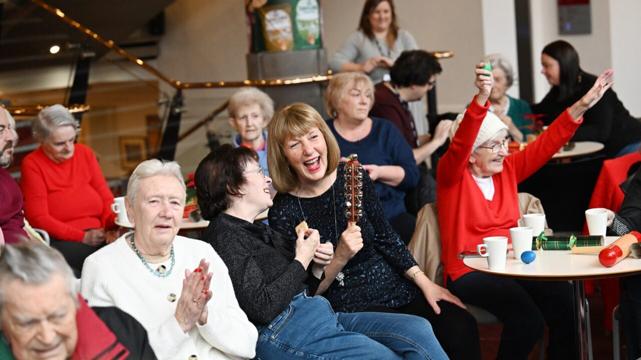 A group of older adults sit together indoors, some holding musical instruments and clapping, while one woman raises her arm enthusiastically; coffee cups are on the tables.