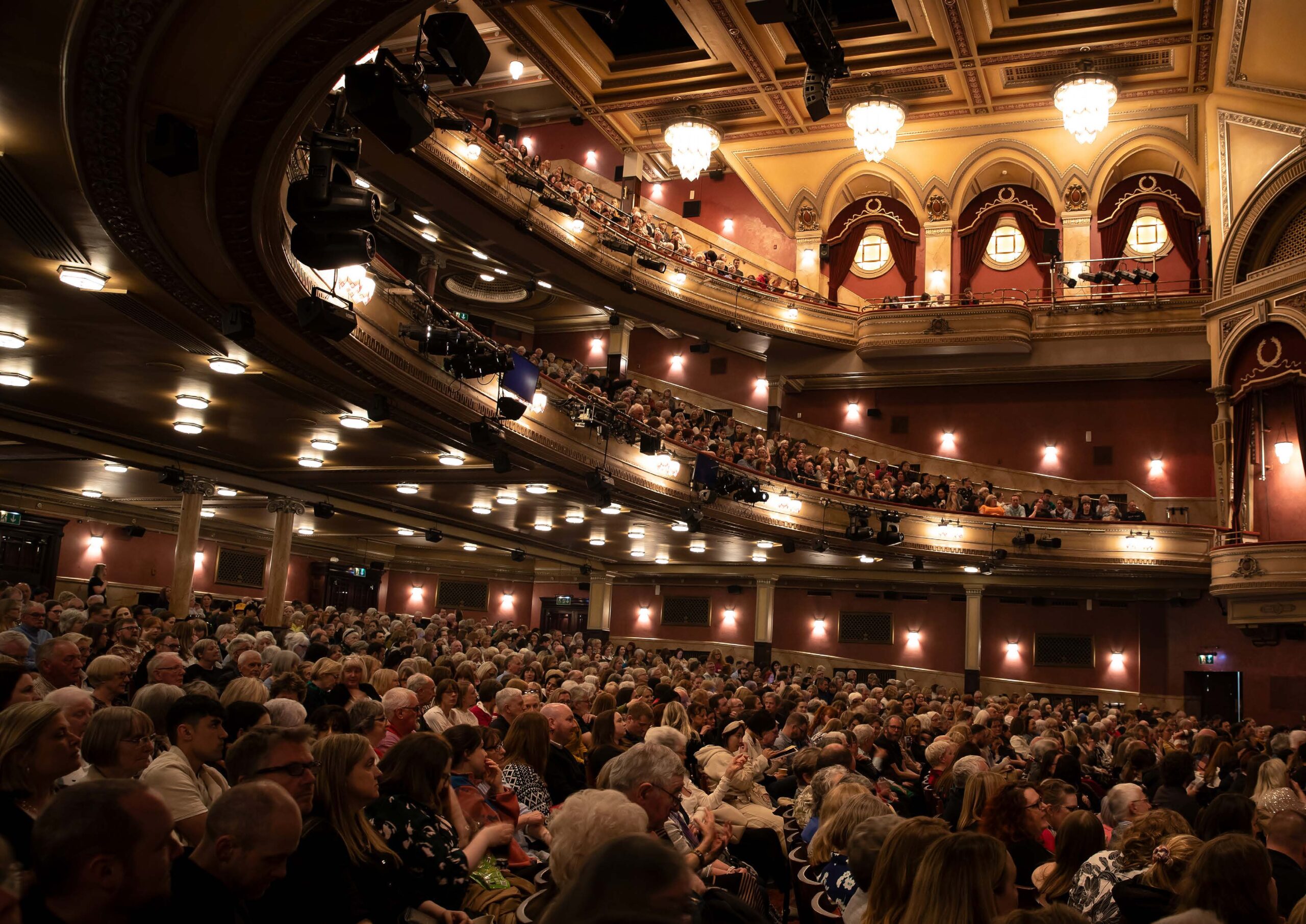 A large audience fills the seats of an ornate, multi-tiered theater, waiting for a performance to begin.