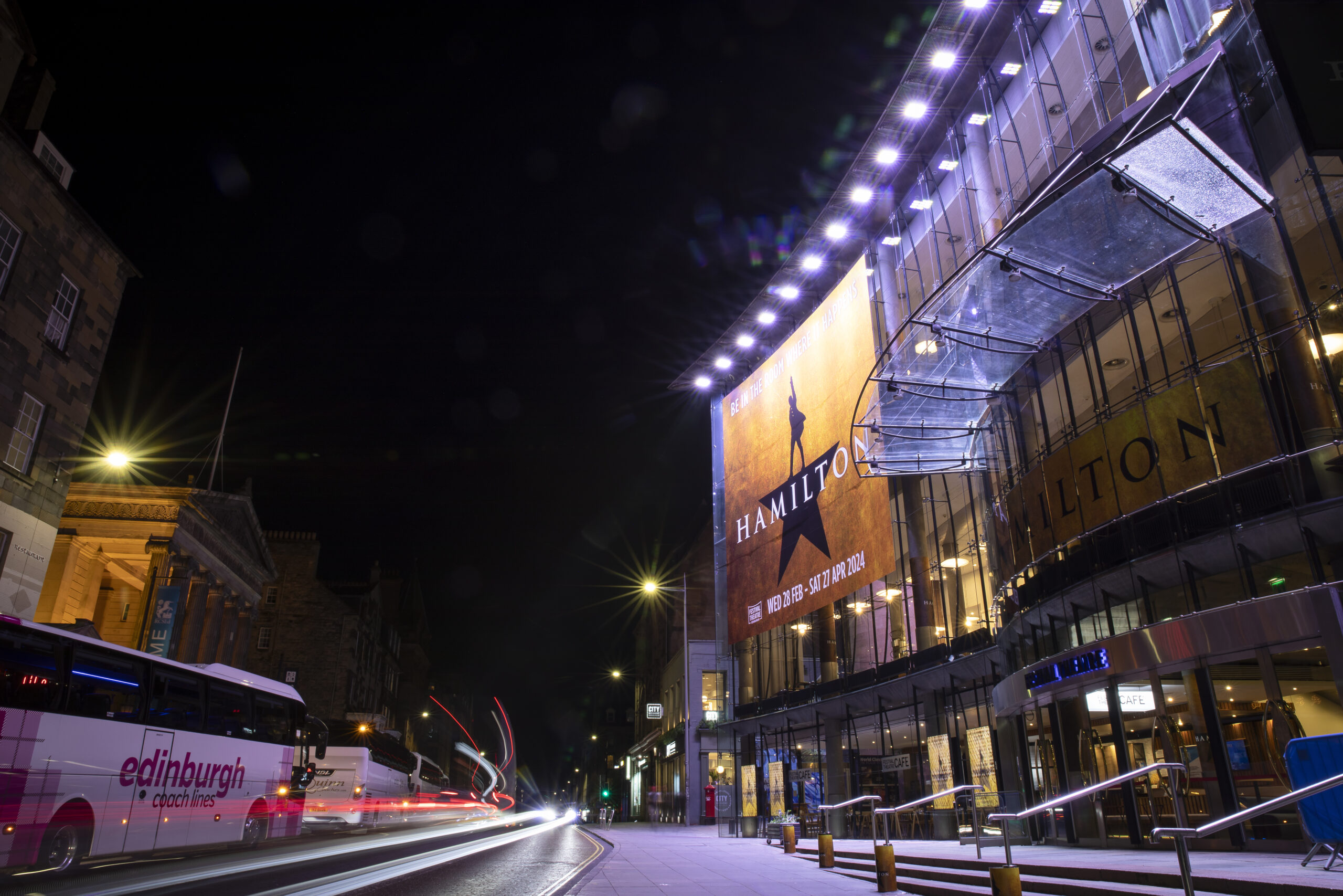 Night view of a theatre with a large "Hamilton" musical poster, illuminated entrance, and a passing Edinburgh city bus on a street with light trails.