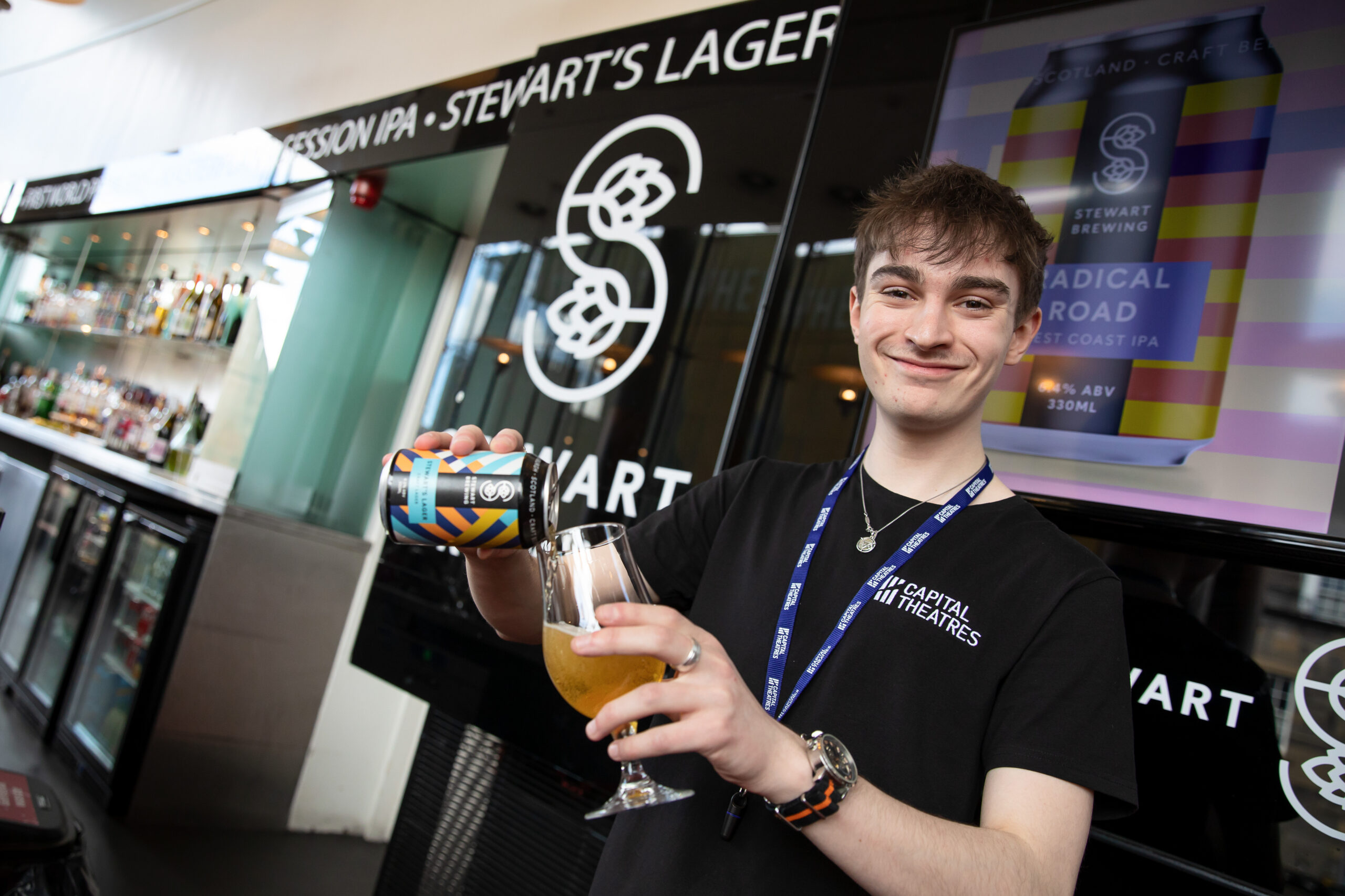 A person in a black t-shirt pours beer from a can into a glass at a bar, with Stewart’s Brewing branding visible in the background.