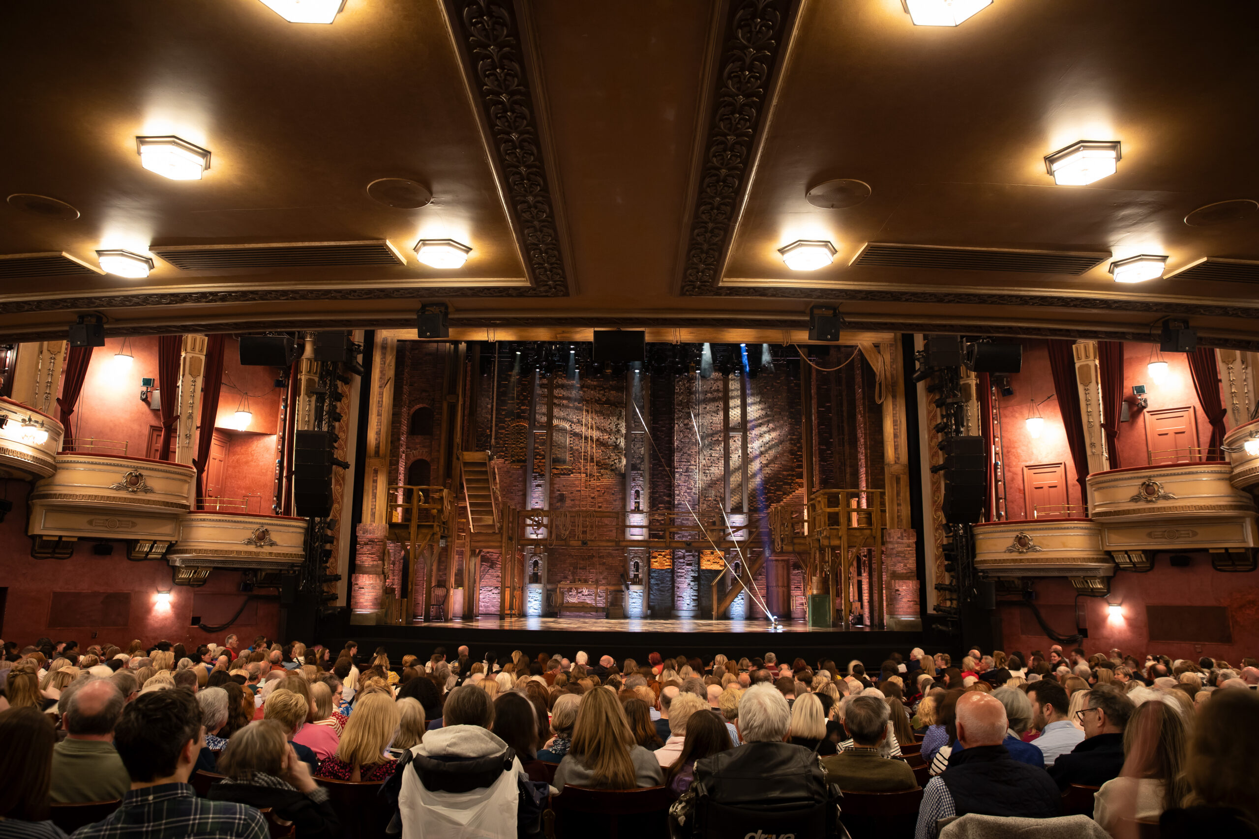 A full audience sits in a historic theatre facing a stage set with wooden scaffolding and brick walls, awaiting the start of a performance.