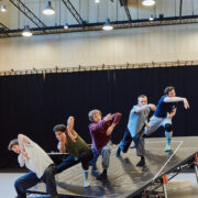 Five dancers perform on an angled platform indoors, each striking expressive, synchronized poses under overhead lights.