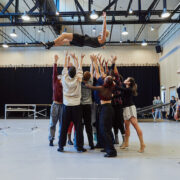 A group of dancers lifts a person high above their heads in a brightly lit studio, while another dancer stands to the side watching.