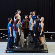 A group of men stands in two facing rows on a raised platform in a rehearsal space with a black curtain backdrop.