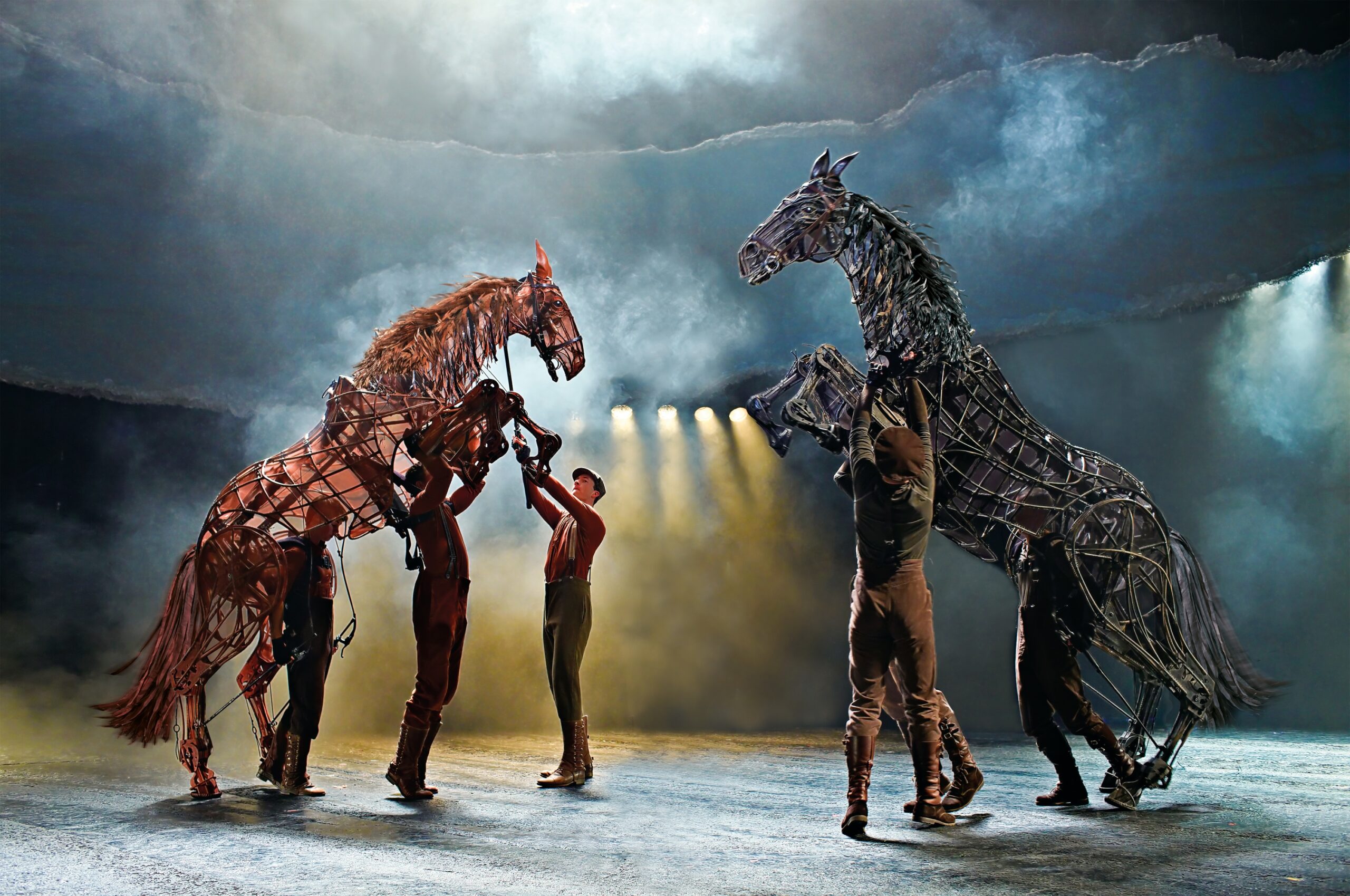 Performers control large, lifelike horse puppets on a misty stage with dramatic lighting and a smoky backdrop.