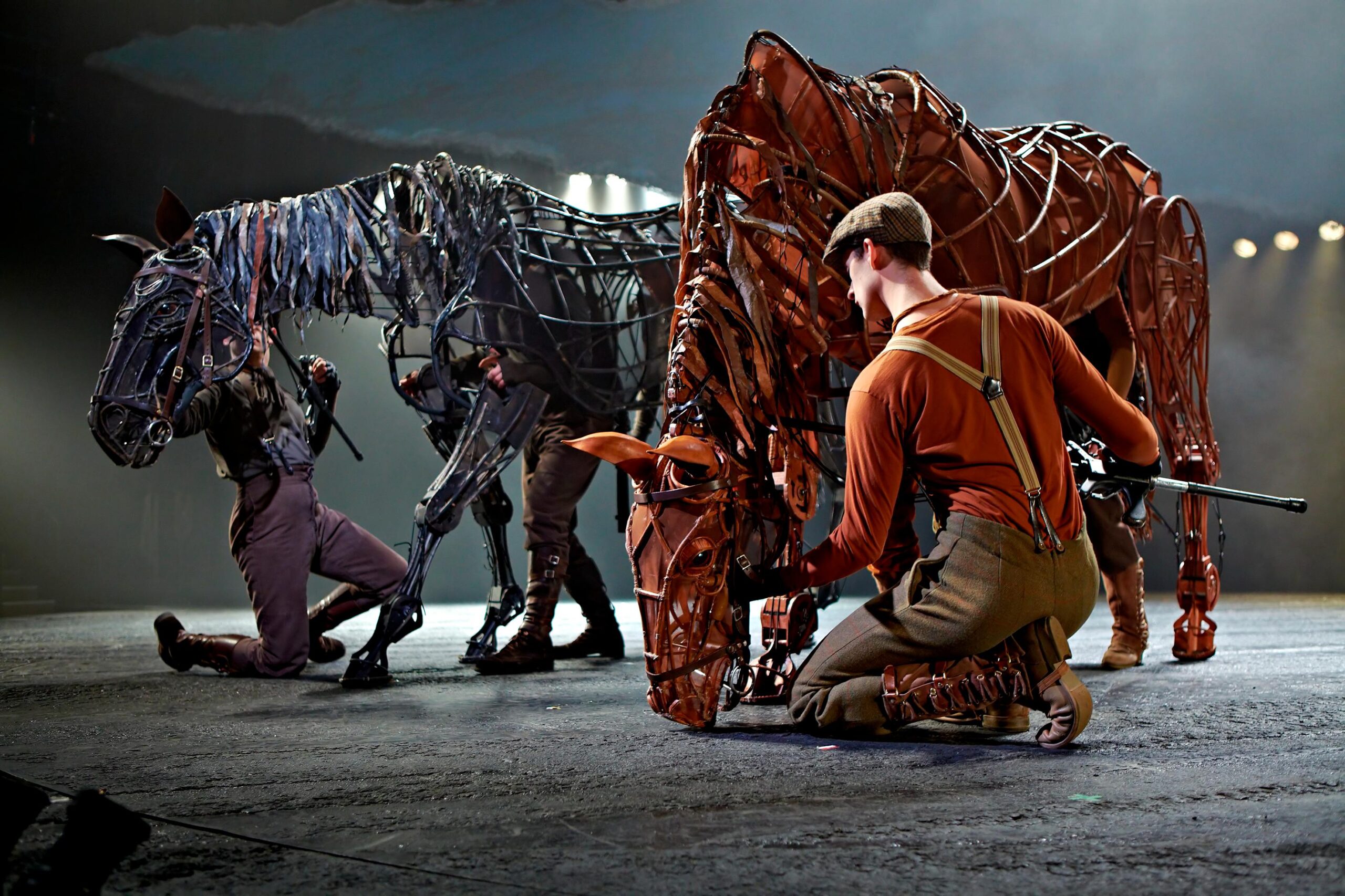 Actors manipulate large, detailed horse puppets on stage during a theatrical performance, with dramatic lighting highlighting the scene.