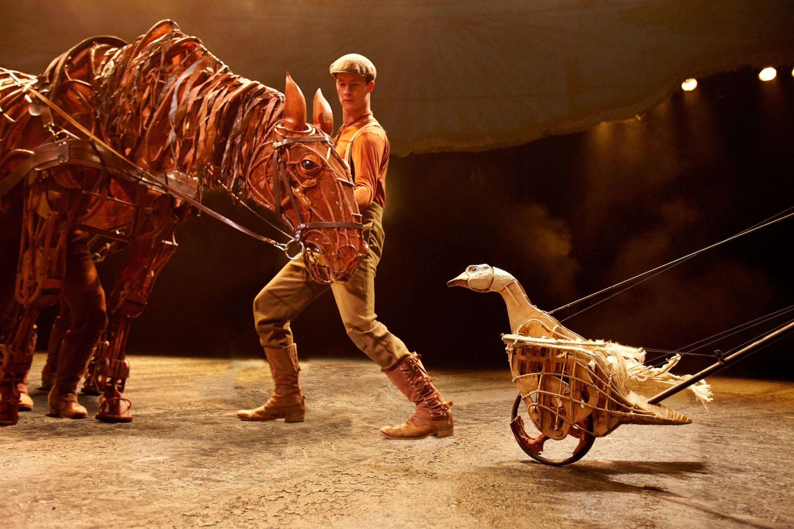 A performer on stage handles a large horse puppet while a goose puppet on wheels, controlled by rods, is positioned nearby under stage lighting.