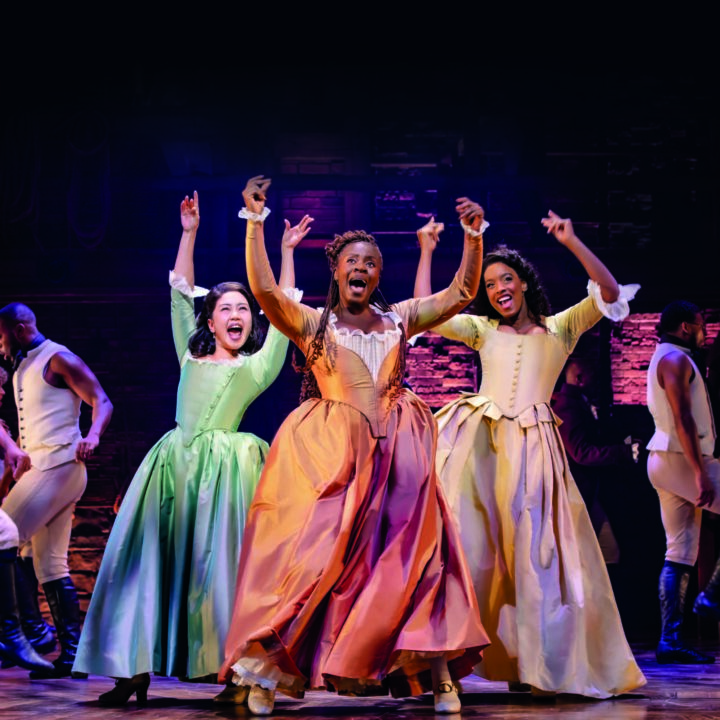 Performers on stage in period costumes, dancing energetically under bright lights.
