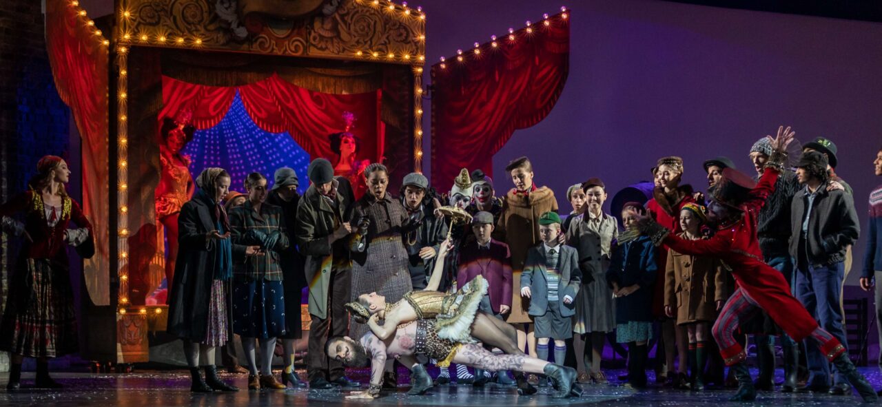 A theatrical stage scene features a large group of actors in period costumes, with two performers dramatically posed at center stage under bright lights.