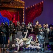 A theatrical stage scene features a large group of actors in period costumes, with two performers dramatically posed at center stage under bright lights.