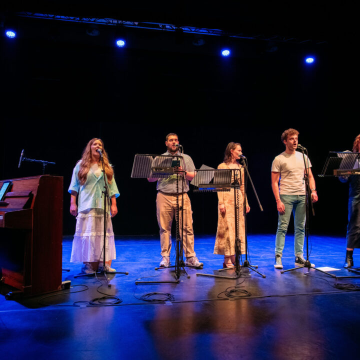 Six performers stand on a stage with microphones; one plays piano while the others sing, under blue stage lights.