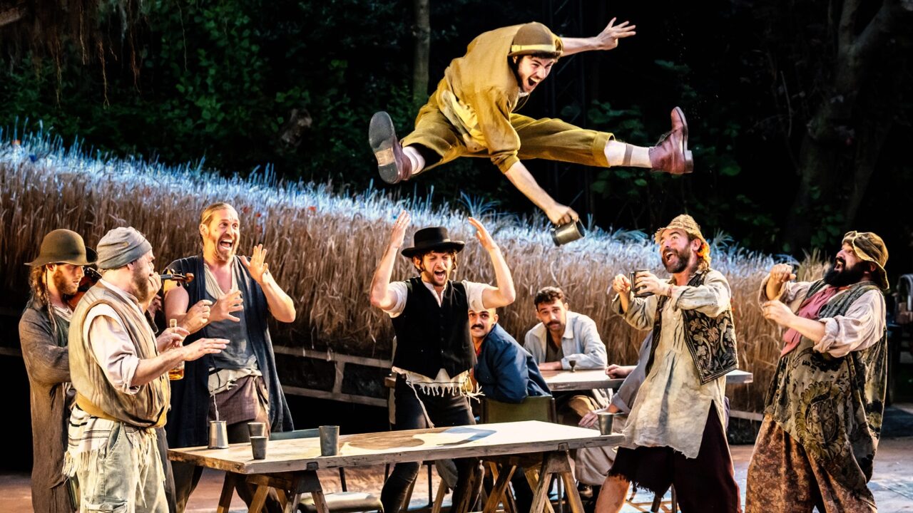 A group of men in period costumes watch as one man leaps high above a table with arms outstretched, mid-performance in an outdoor theatrical play.