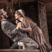 Two actors in period costumes hold hands and smile at each other while kneeling on a wooden table during a stage performance.