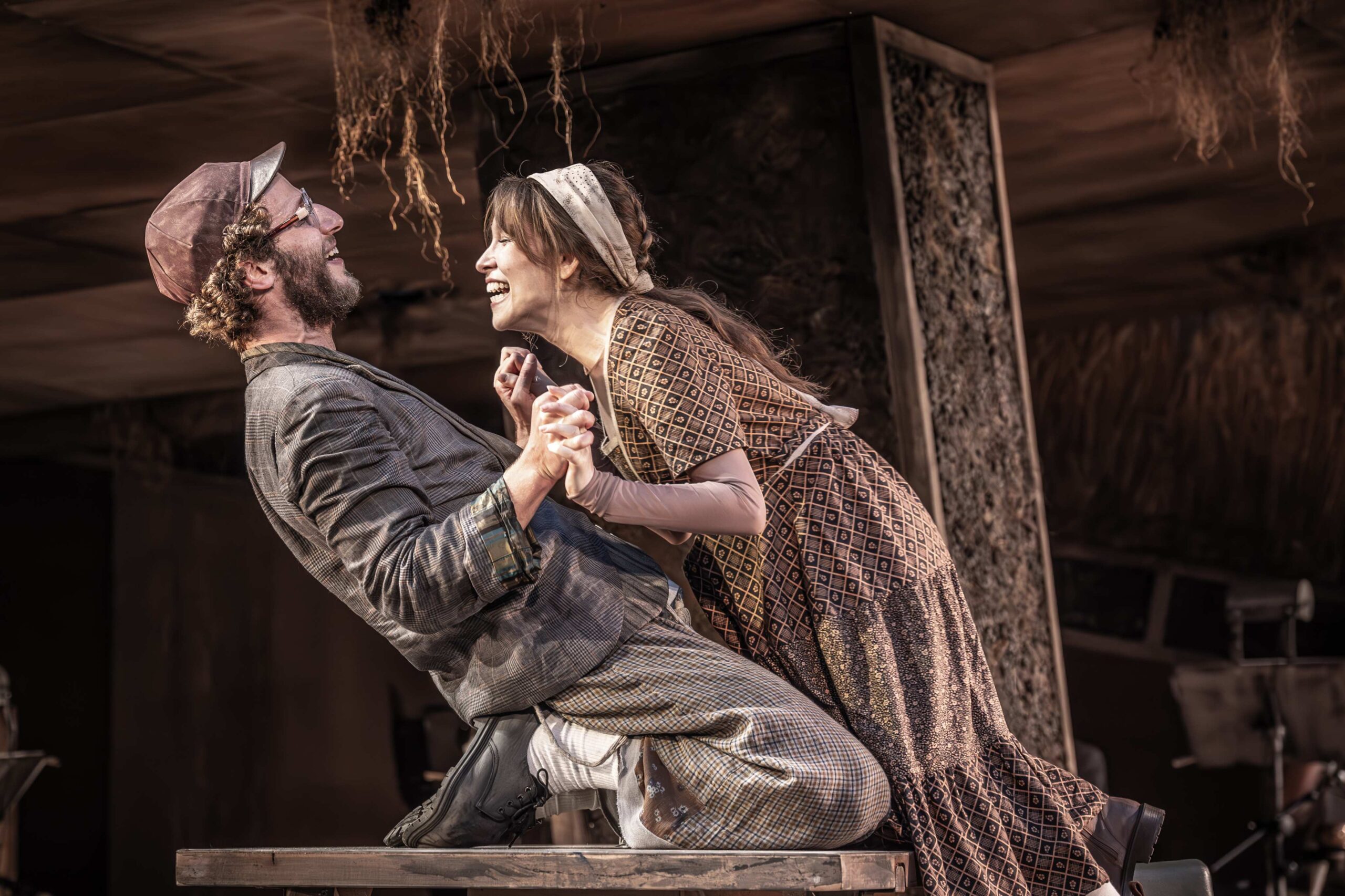 Two actors in period costumes hold hands and smile at each other while kneeling on a wooden table during a stage performance.