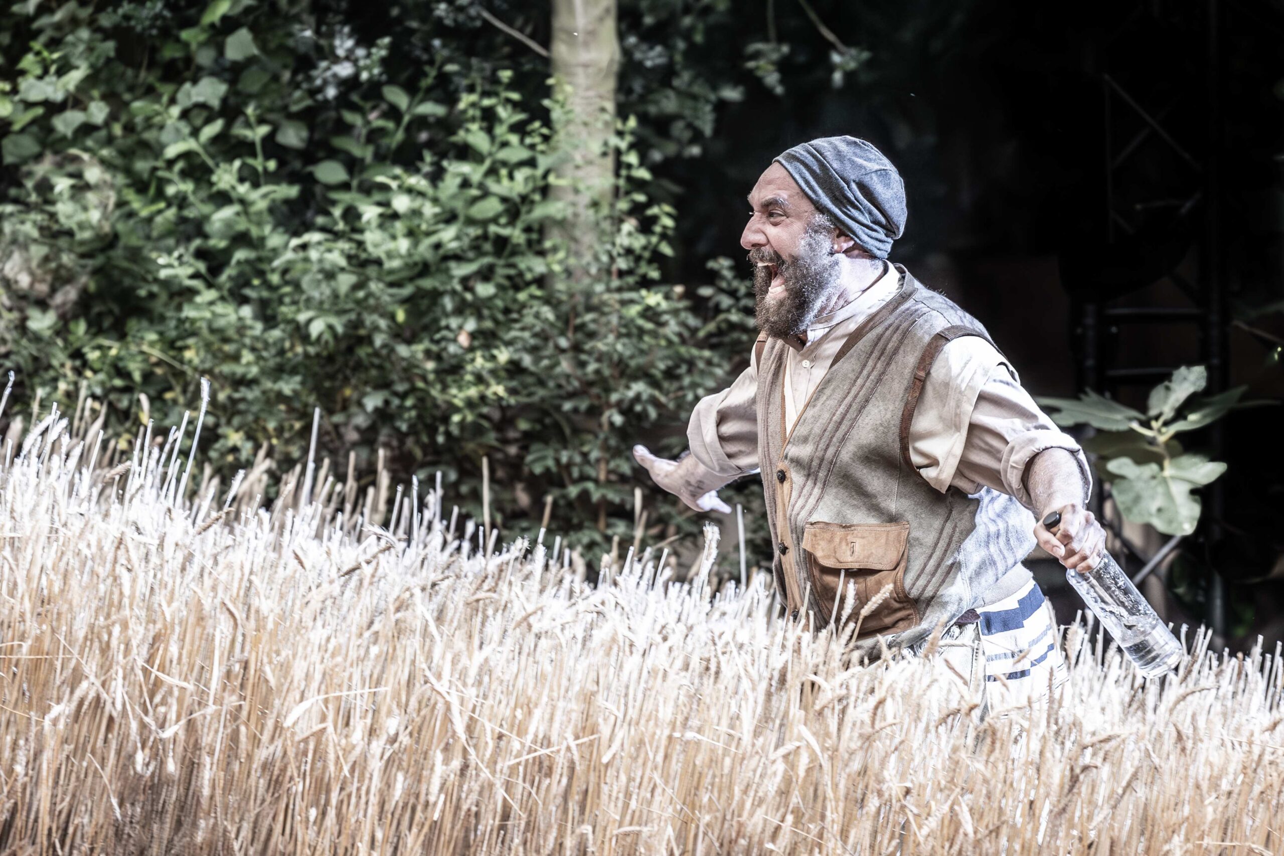 A bearded man in rustic clothing stands in a wheat field with arms outstretched, smiling. Dense green foliage is visible in the background.