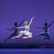 A ballerina in a white tutu performs a mid-air leap, flanked by two other dancers in blue costumes, against a purple background with silhouette trees.