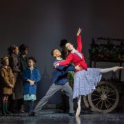 A group of people watch as a male and female ballet dancer perform in front of a flower cart on stage, with the woman posed en pointe and the man supporting her.