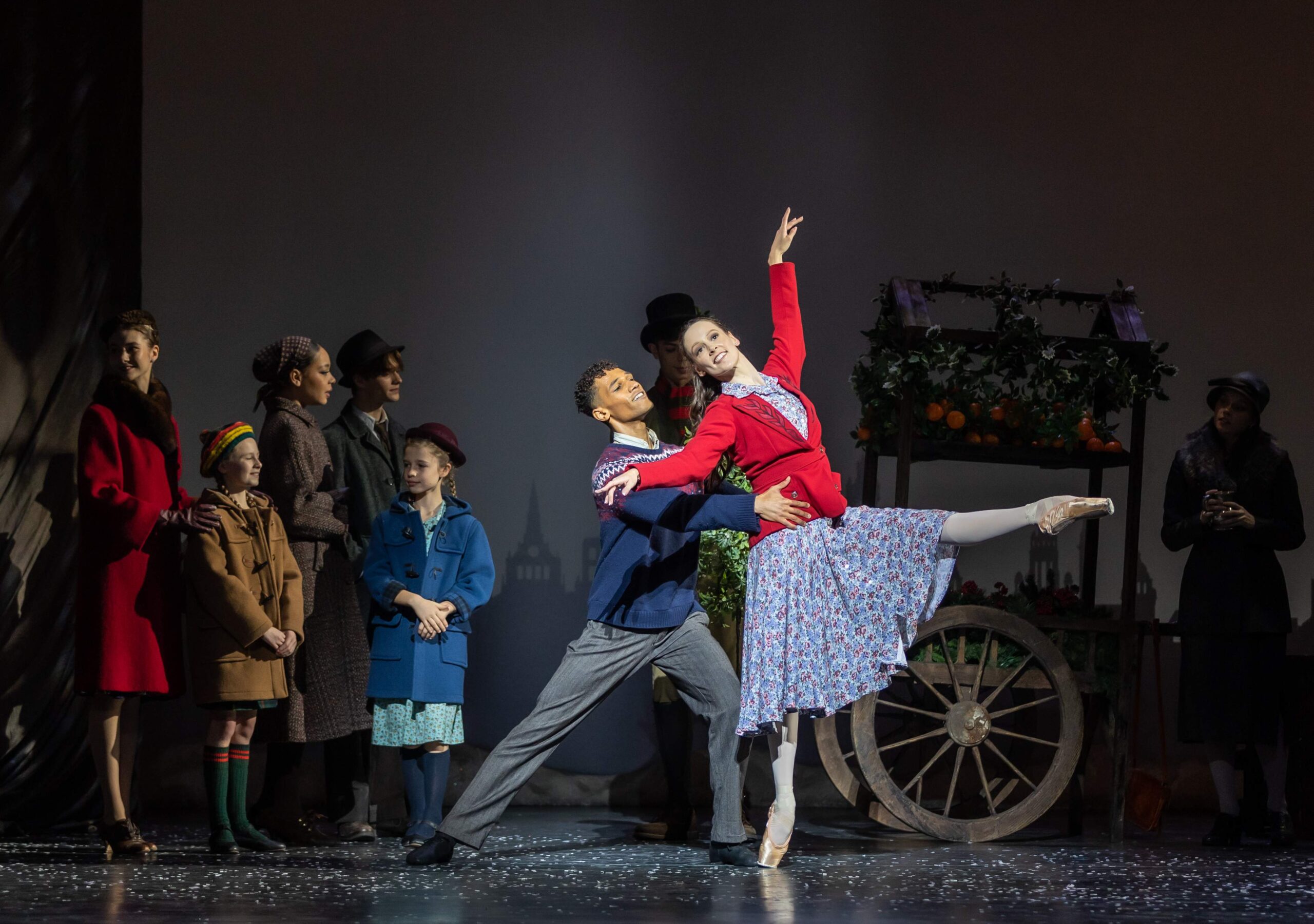A group of people watch as a male and female ballet dancer perform in front of a flower cart on stage, with the woman posed en pointe and the man supporting her.