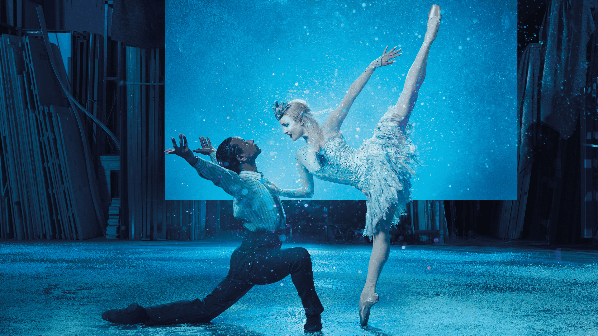 Two ballet dancers perform on stage; one is kneeling and reaching upward while the other balances on pointe with one leg extended, surrounded by blue lighting and a snowy effect.