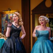Two women in vintage-style dresses, one in black and one in blue, stand side by side indoors, smiling and raising their gloved hands as if in mid-conversation or greeting.