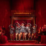 A group of performers in glittering costumes and top hats dance on stage under red lighting, surrounded by an audience seated at tables.