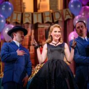 Three people in vintage clothing perform on stage at a birthday-themed set with balloons and a "BIRTHDAY" banner in the background.