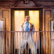 A man in striped pajamas stands surprised on a balcony in front of glass doors, with a statue of the Virgin Mary holding baby Jesus on a pedestal nearby.
