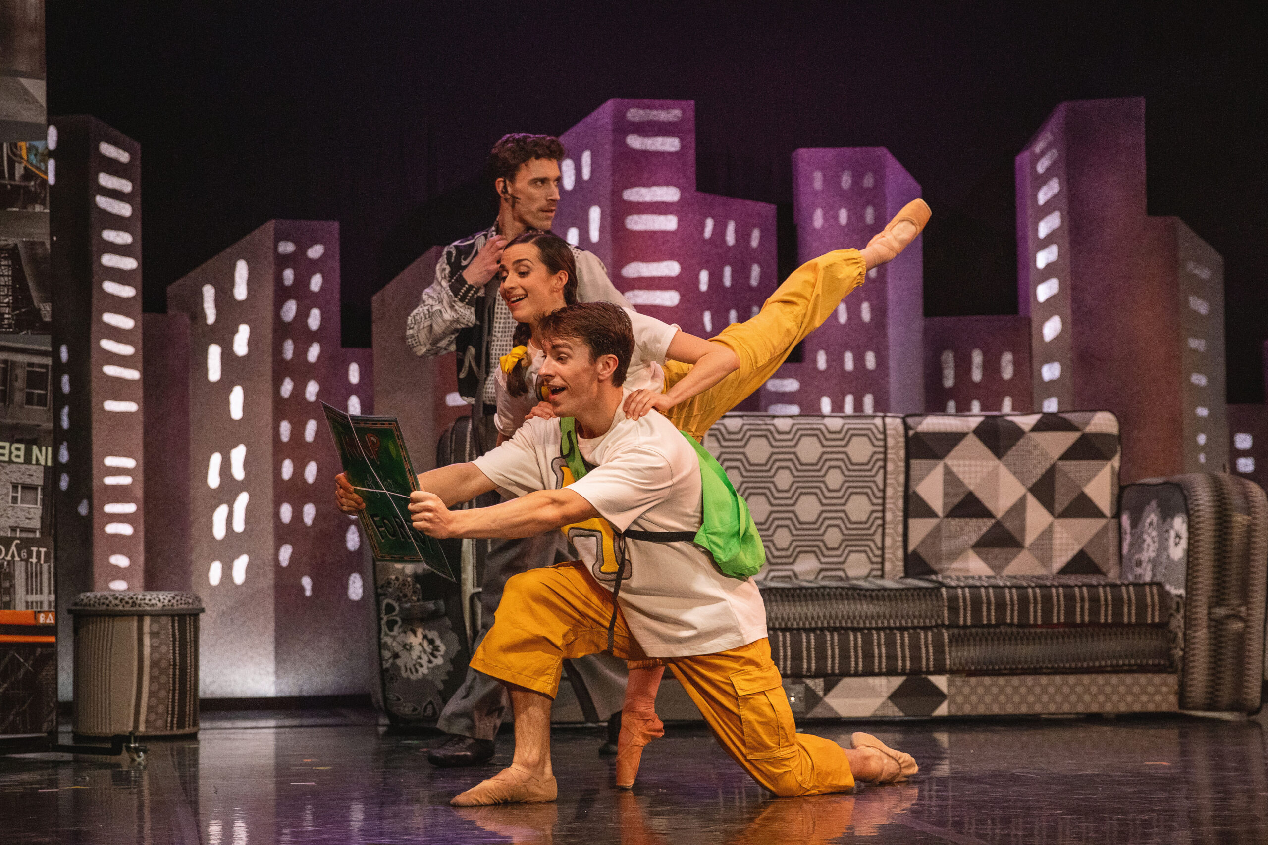 Three performers in colourful costumes pose energetically on stage with a cityscape backdrop, one holding a framed picture, while another balances on one leg behind him.