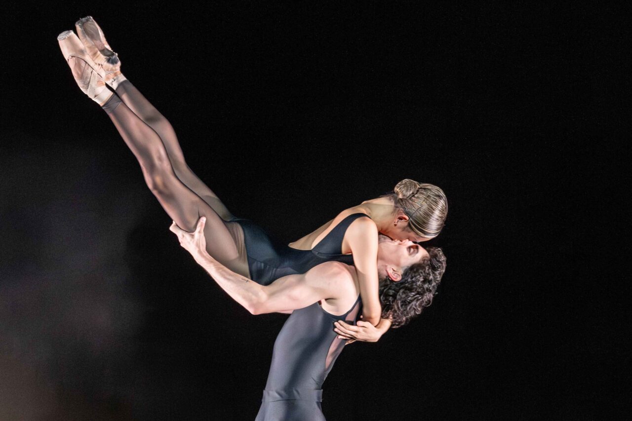 A male ballet dancer in a black outfit lifts a female dancer in a black leotard and pointe shoes horizontally above his head against a dark background.