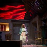 A woman stands center stage in a dimly lit, damaged room with red clouds projected behind her, wearing a cardigan and long skirt, her hands held in front of her.