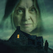 A stone house with one lit window sits under a dark, cloudy sky. A large, transparent face of an older woman looms above, while a lone figure stands outside.