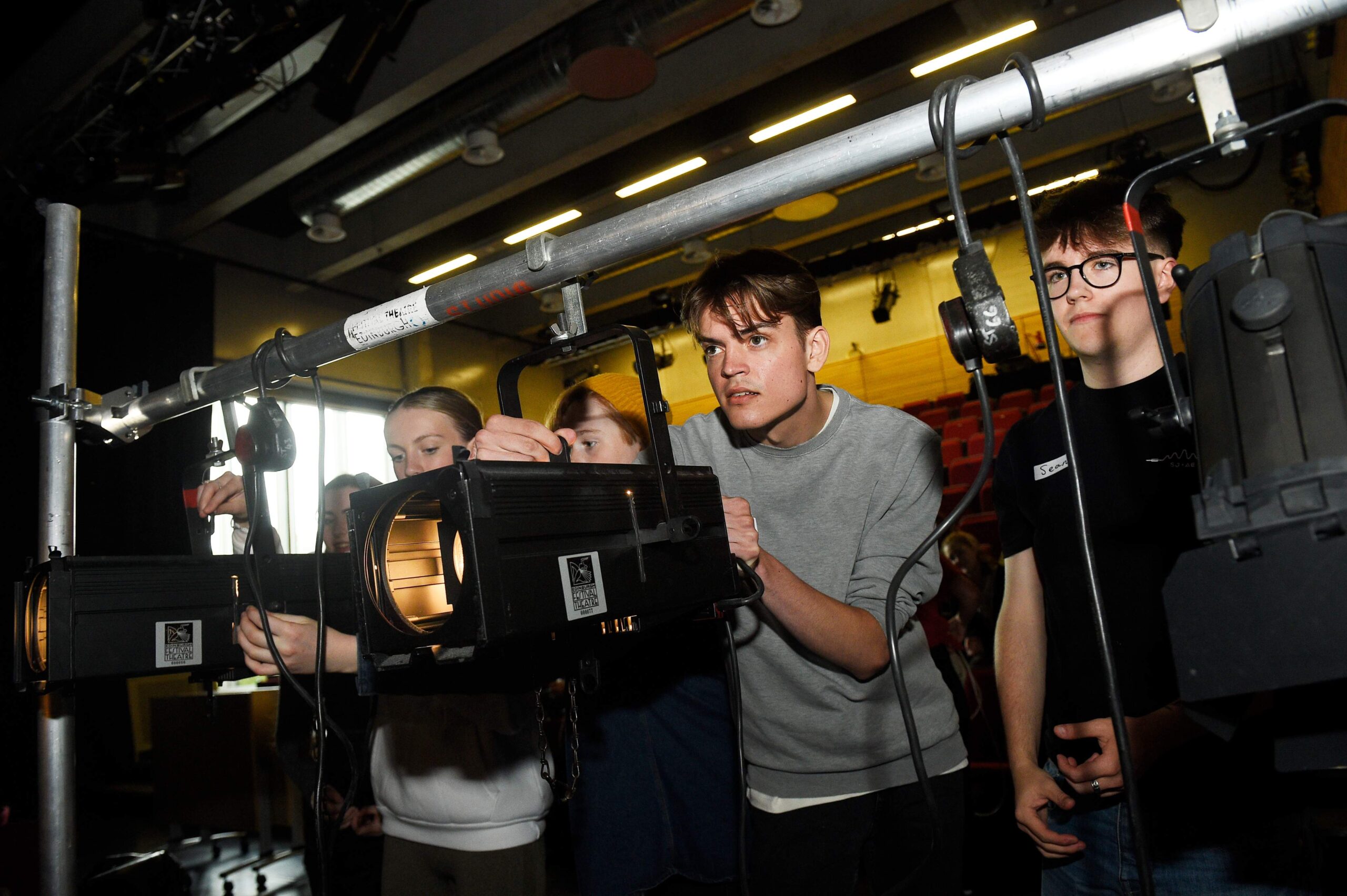 A group of people adjusting stage lighting equipment, focusing on positioning light fixtures on a metal rig in a theatre setting.