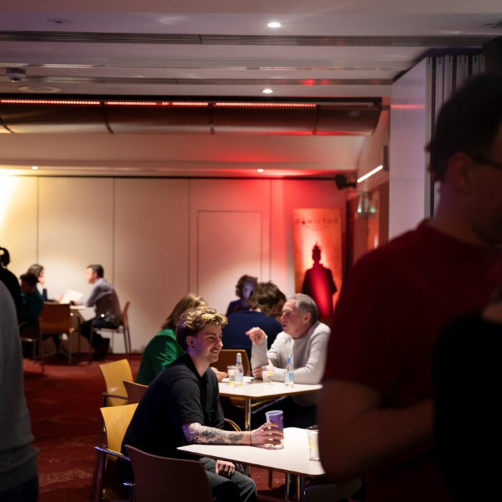 People sit and talk at tables in an atmospherically lit room with red carpeting and warm lighting on the walls. Some individuals stand and converse in the foreground.