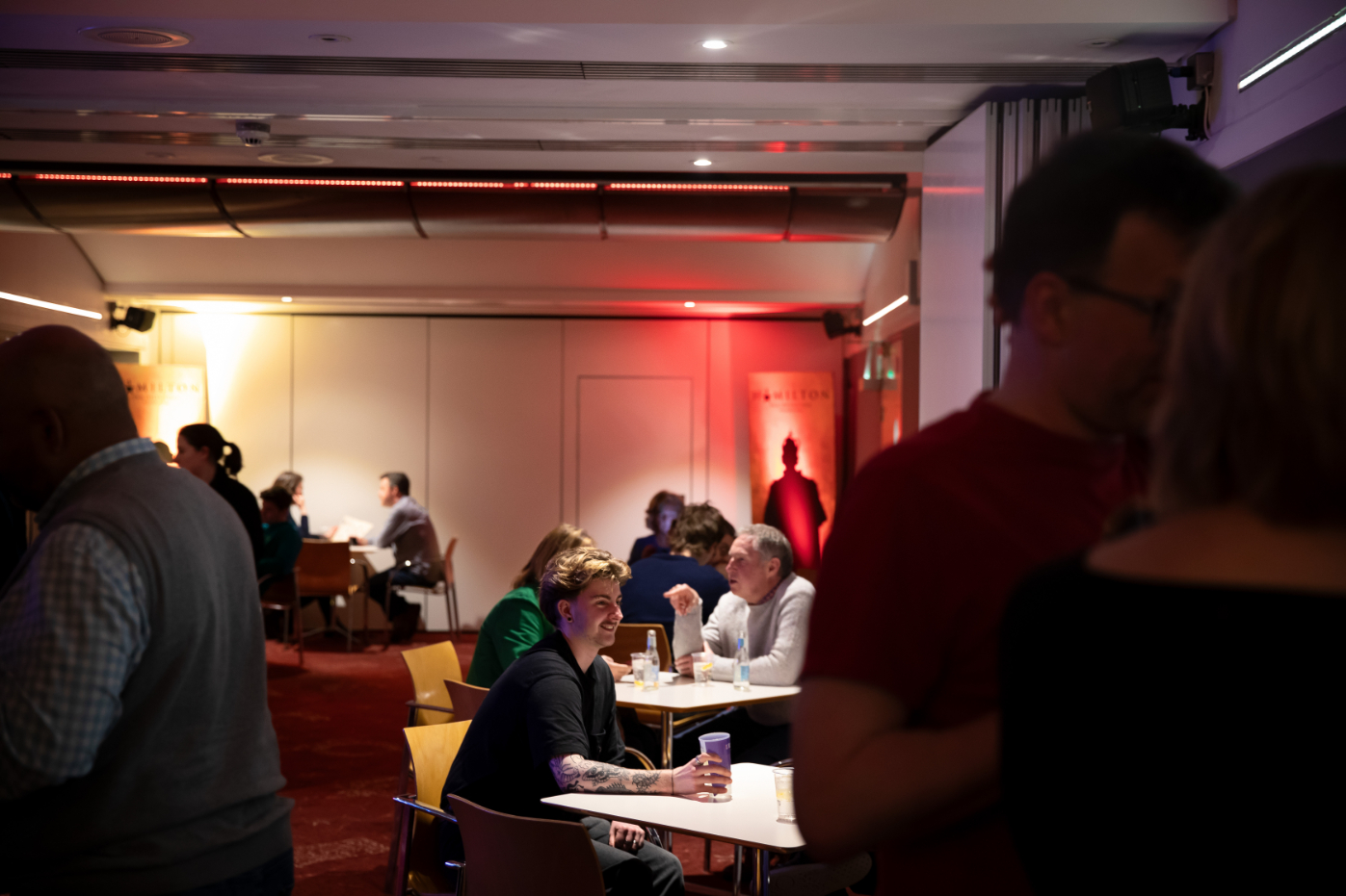 People sit and talk at tables in an atmospherically lit room with red carpeting and warm lighting on the walls. Some individuals stand and converse in the foreground.