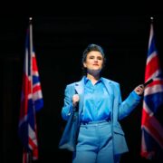 A person in a blue suit stands on stage between two British flags, holding a pen and looking ahead under bright stage lighting.